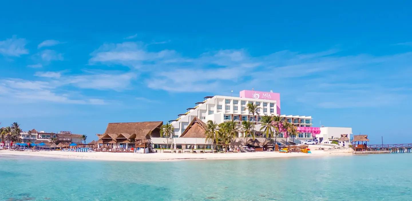 Beachfront exterior of Mia Reef Isla Mujeres with turquoise water, palms, and thatched palapas.