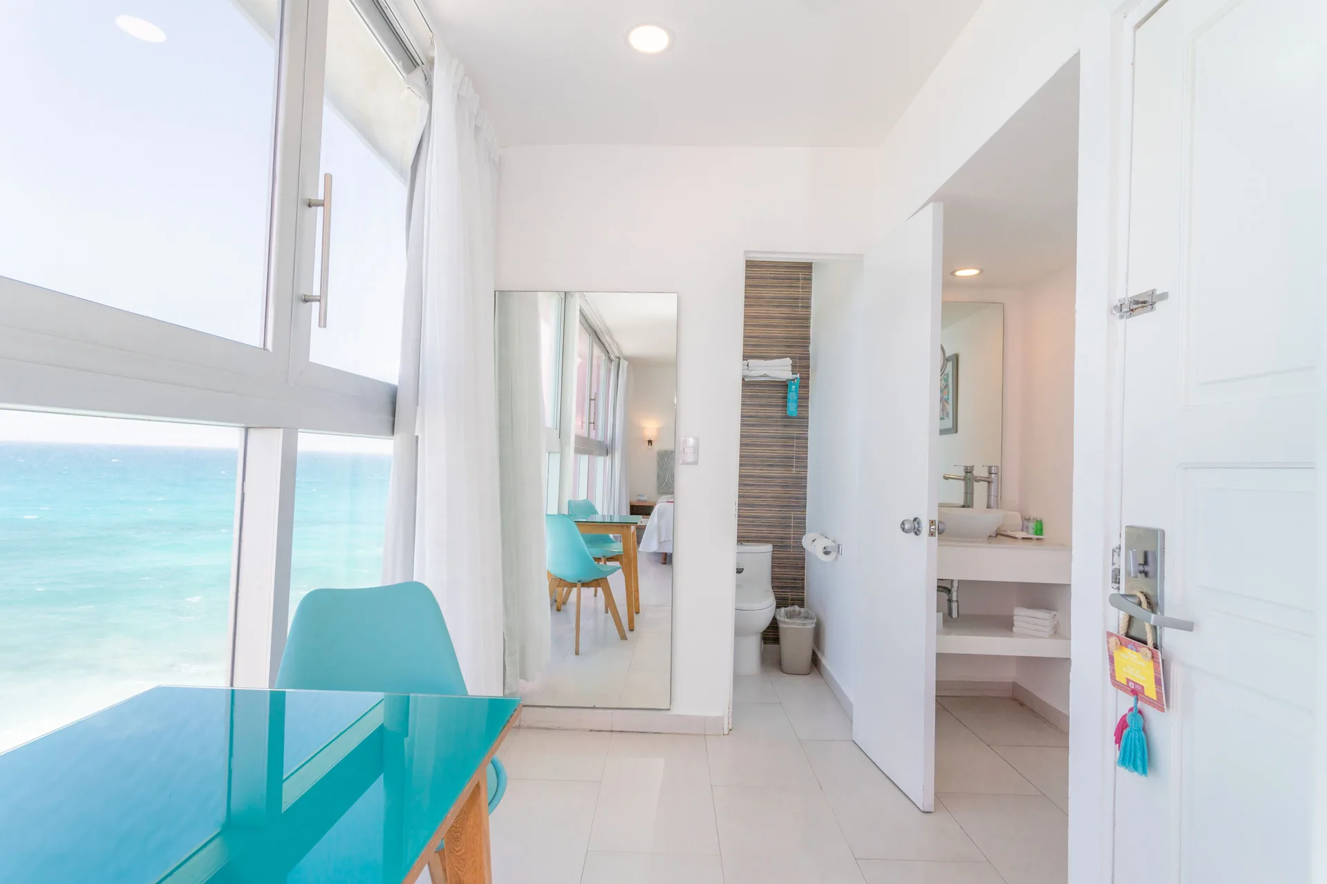 Guest room entry at Mia Reef Isla Mujeres with turquoise desk, chair, ocean window, and bathroom doorway.