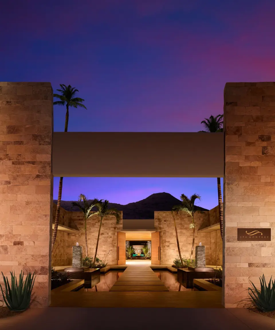 Resort entrance with stone walls and palm trees at dusk at Montage Los Cabos.
