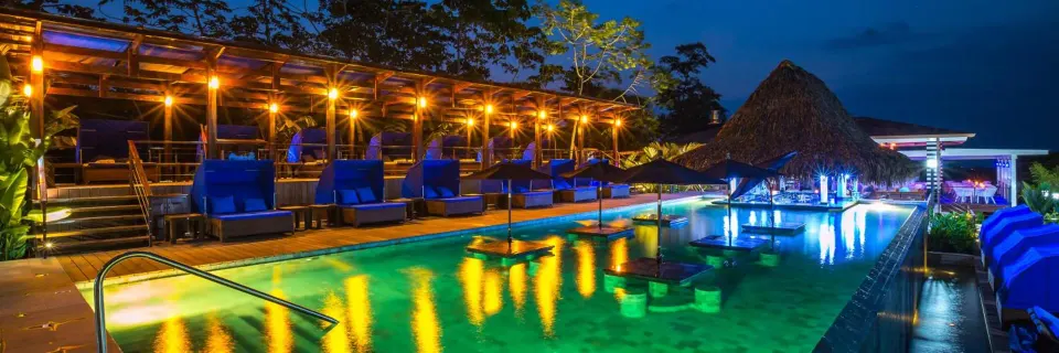 Illuminated resort pool with lounge chairs and a thatched bar at Nayara Springs.