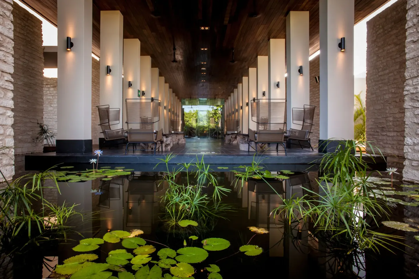 Covered lobby walkway with lily pond, columns, chairs, and stone walls at Nizuc Resort and Spa.