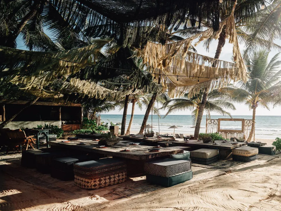 Beach lounge with low tables and poufs under fringed canopies at Nômade Temple Tulum