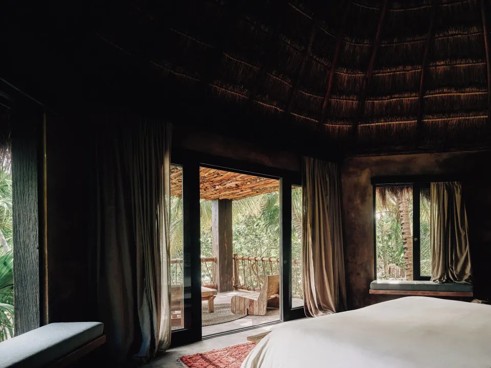 Large king bed in a palapa-ceilinged suite with doors opening to a covered balcony at Nômade Temple Tulum