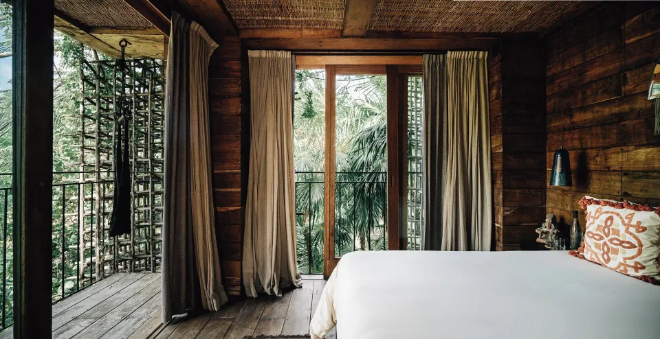 Wood-paneled guest room with king bed opening to a balcony and tropical trees at Nômade Temple Tulum