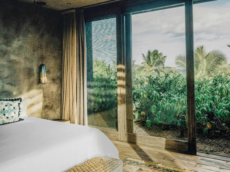 Corner view of the king bed and glass doors overlooking garden at Nômade Temple Tulum