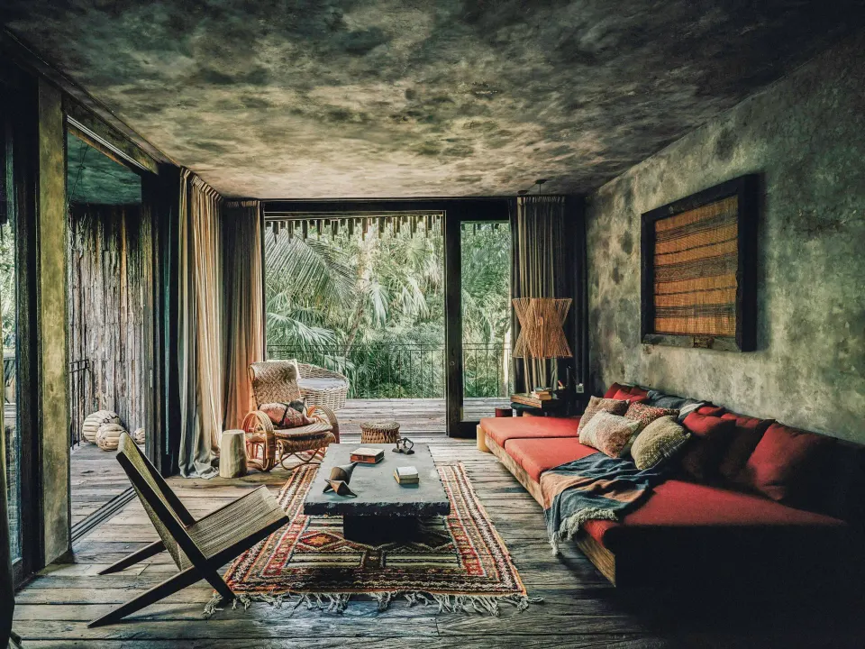 Living area with low red sofa and sliding glass doors opening to jungle in a Deluxe Room at Nômade Temple Tulum