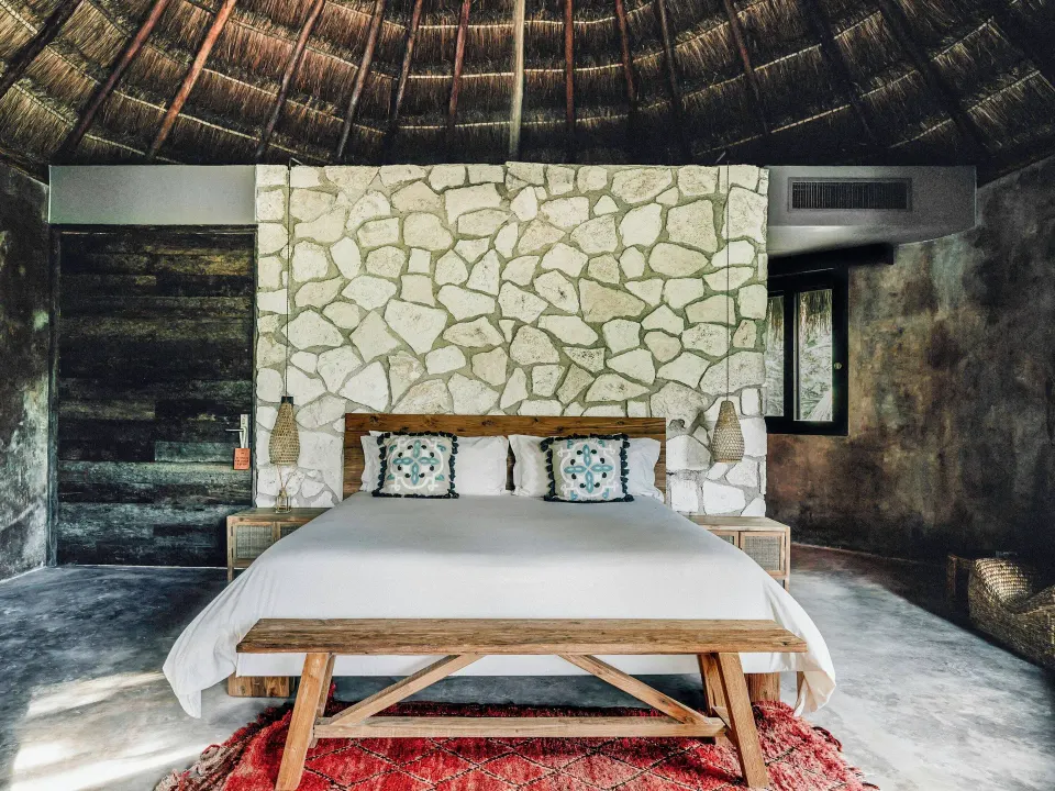 King bed with stone feature wall beneath a palapa ceiling in a Superior Room Beach View at Nômade Temple Tulum