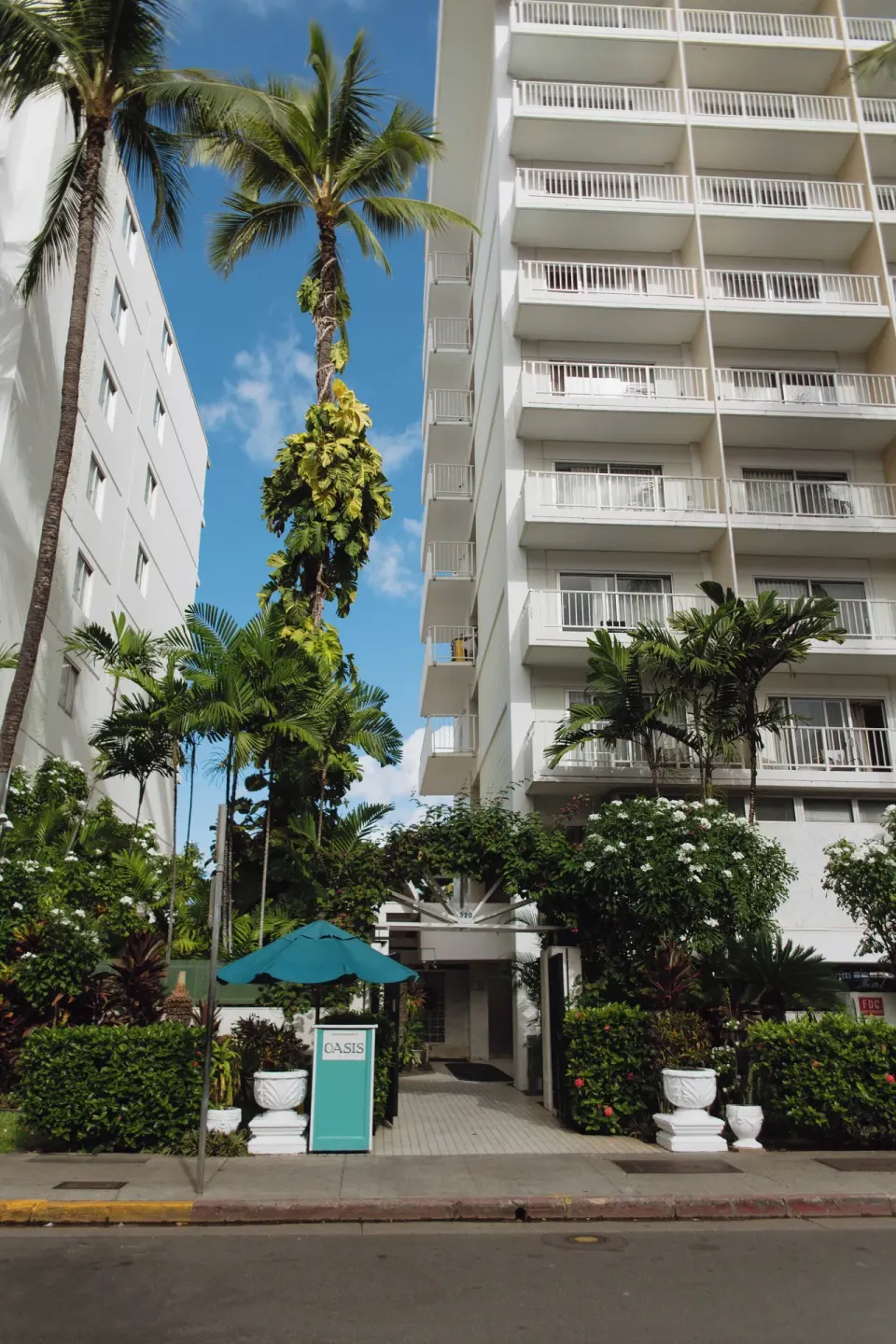 Street entrance with palms, hedges, and a turquoise sign at Oasis Hotel Waikiki.