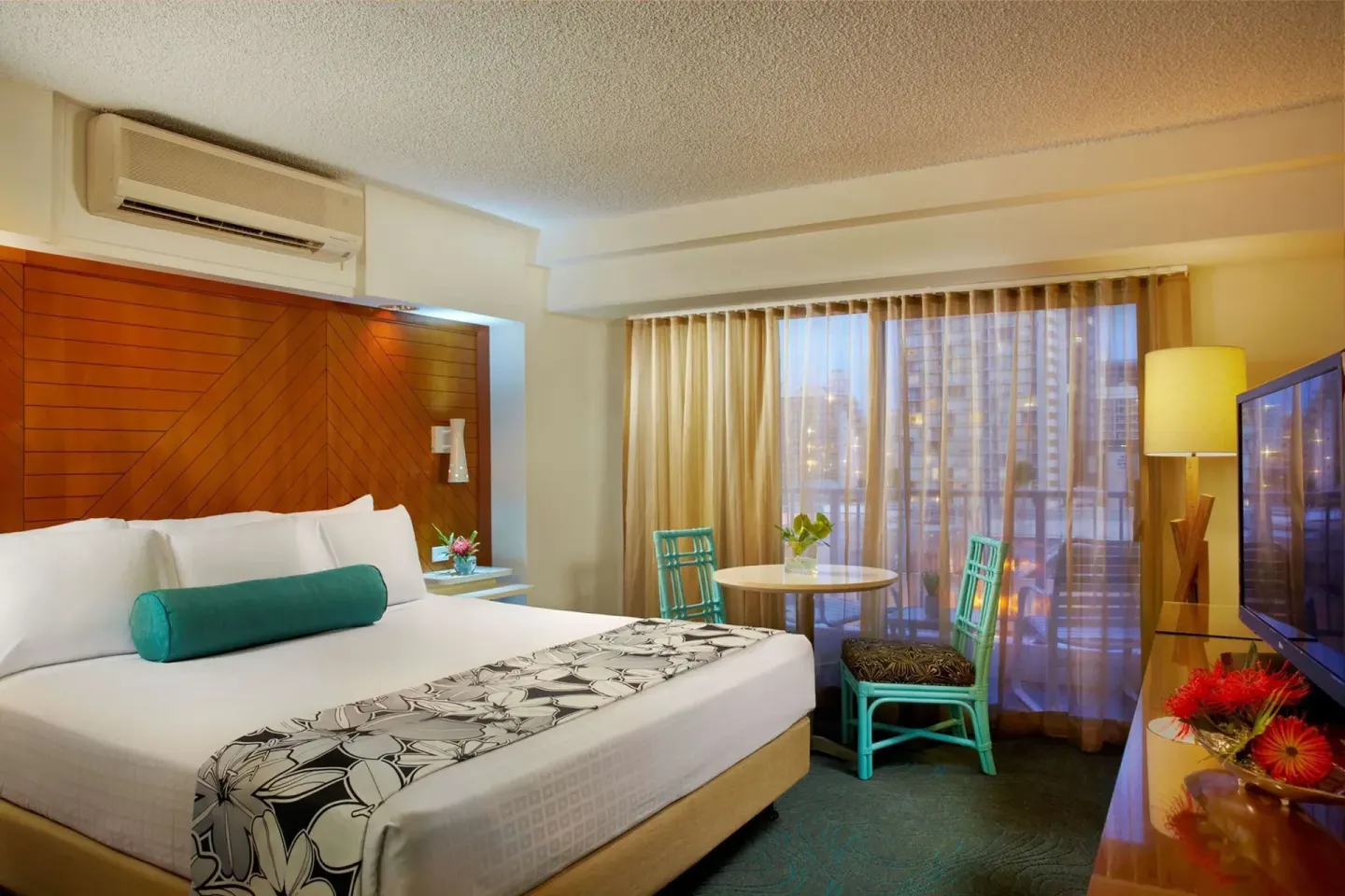 Standard Room king bed beside a round table and curtained window at Oasis Hotel Waikiki.