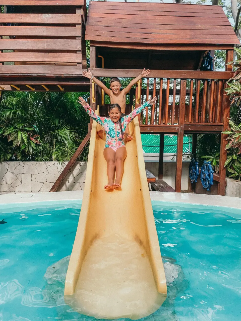 Children ride a yellow slide into a pool below a wooden play structure at Oasis Palm.