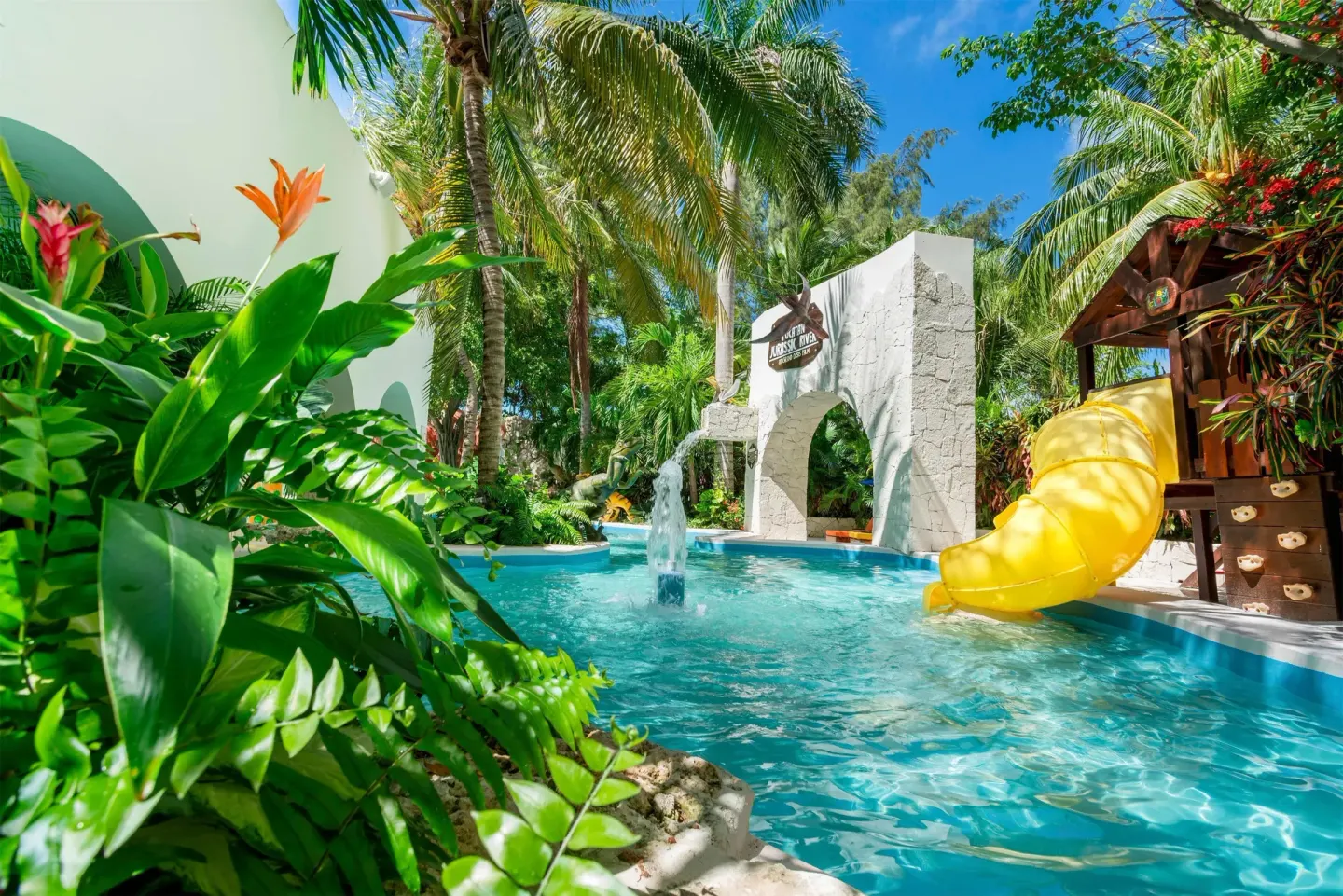 Kids' pool with yellow slide, stone arch, and tropical plants at Oasis Palm.