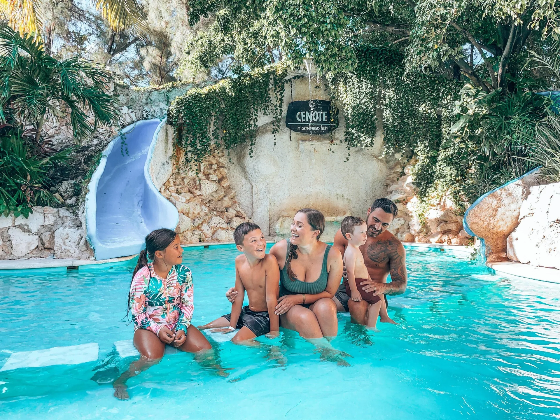 Family poses in a turquoise pool between blue slides and leafy rockwork at Oasis Palm.