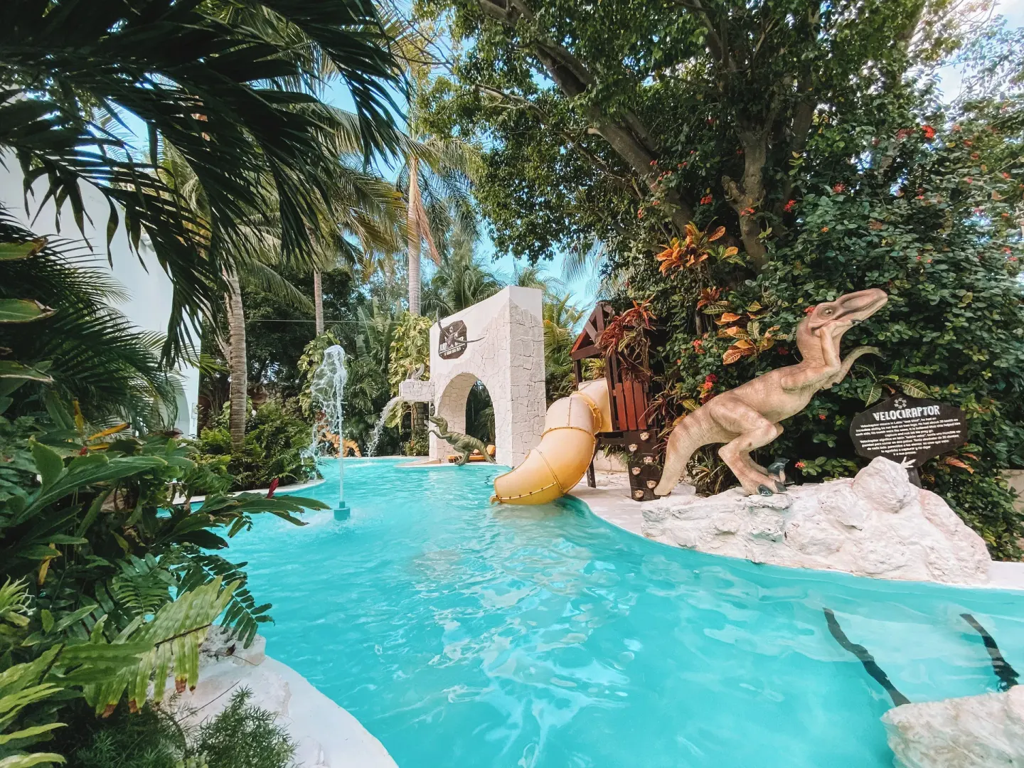 Tropical kids' pool with dinosaur statue, yellow slide, and greenery at Oasis Palm.