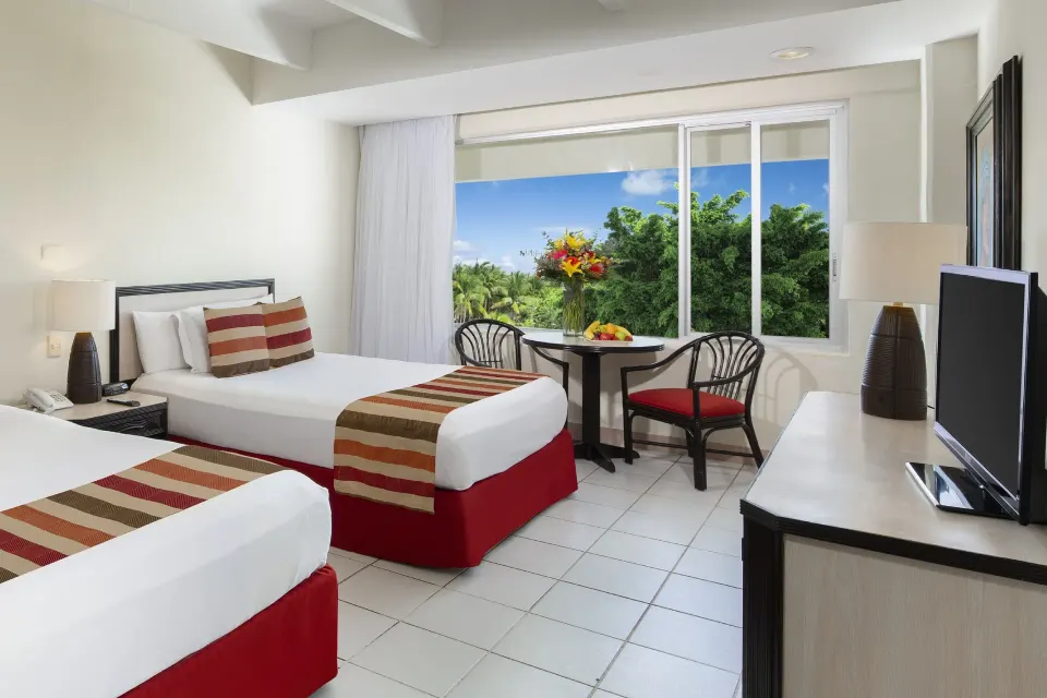 Family room at Oasis Palm with two double beds, a table by the window, TV, and leafy views outside.
