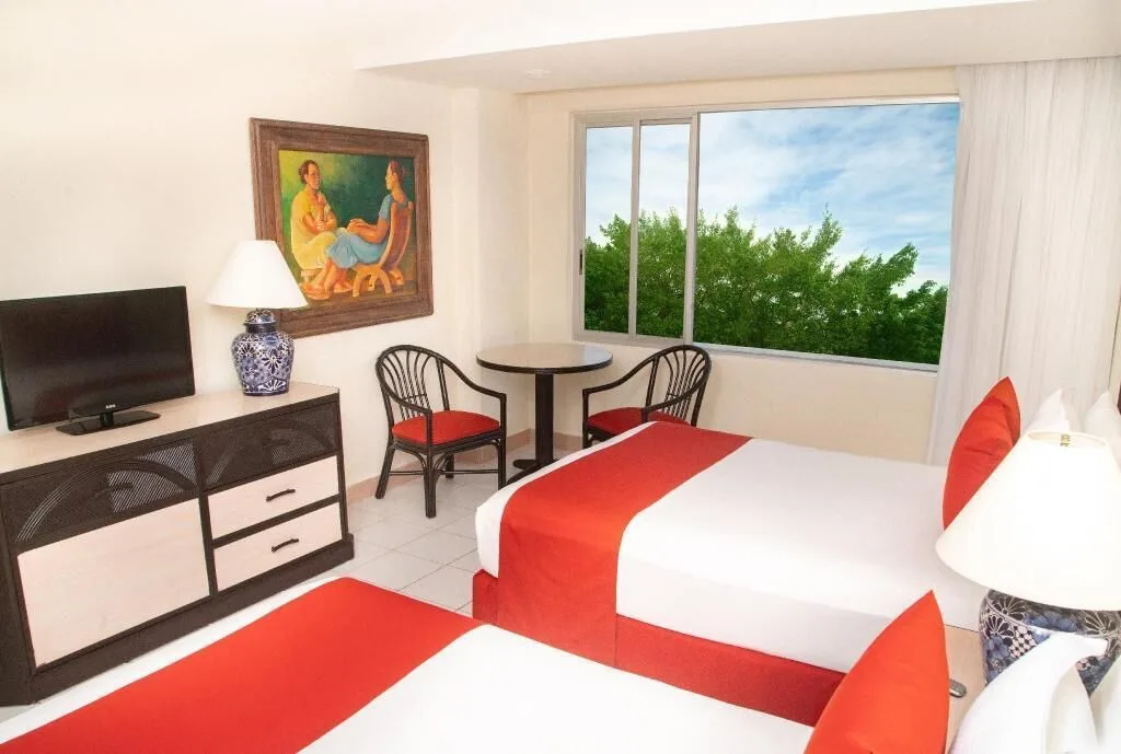 Standard Room at Oasis Palm with two beds, red accents, TV, dining table, and a leafy window view.
