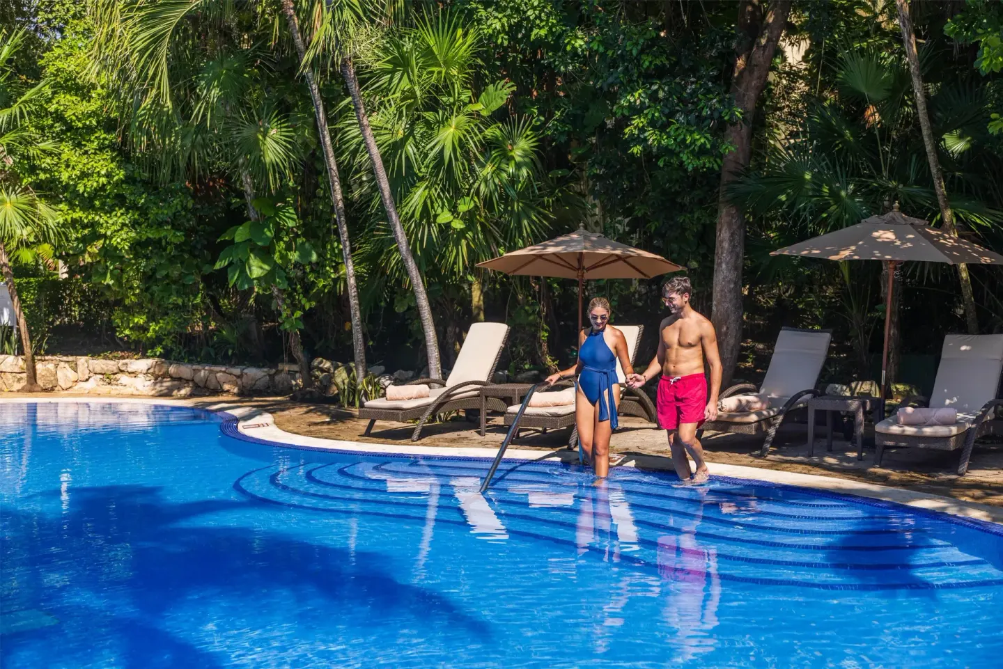 A couple enters a tropical pool beside loungers at Occidental at Xcaret Destination.