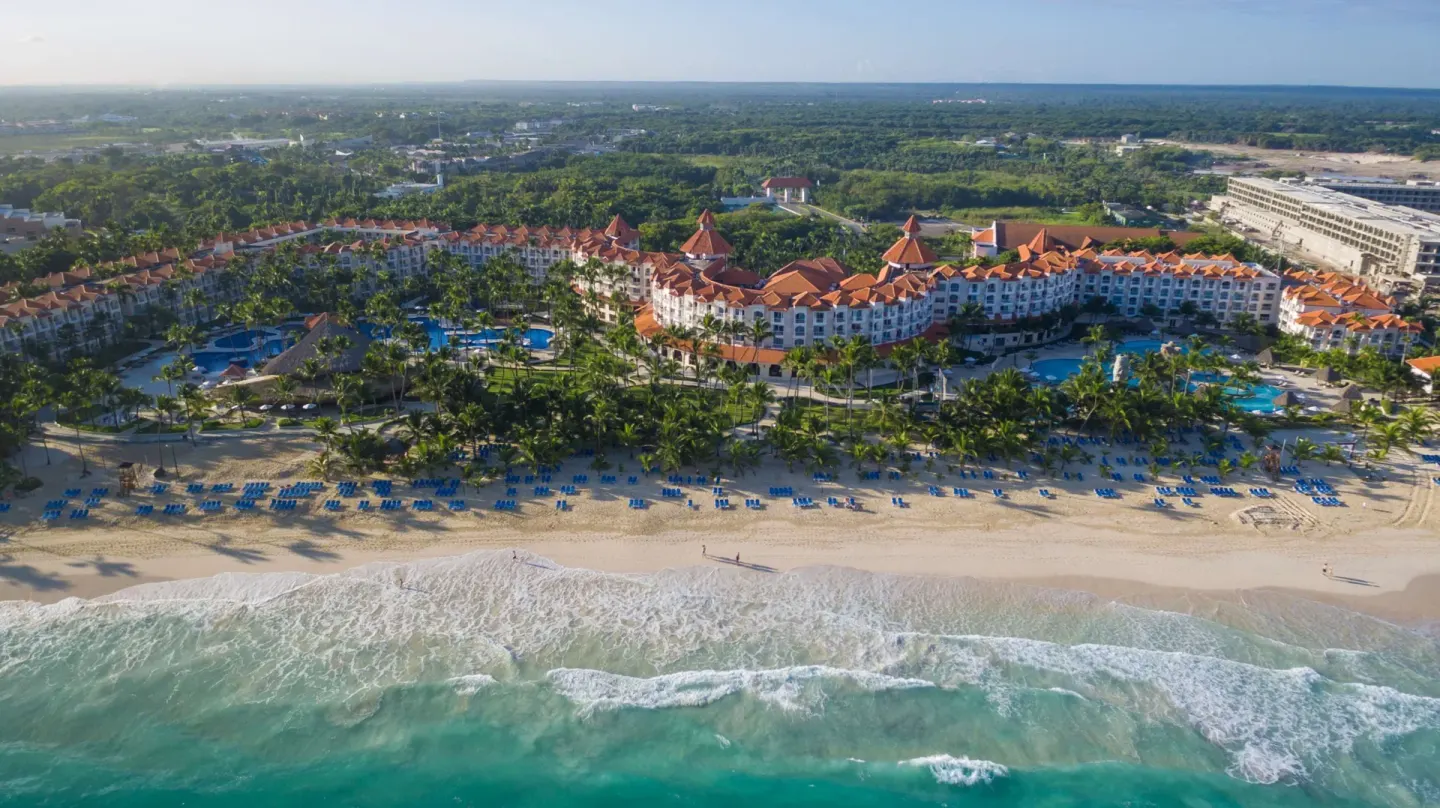 Aerial view shows Occidental Caribe, pools, palms, beach loungers, and ocean surf.
