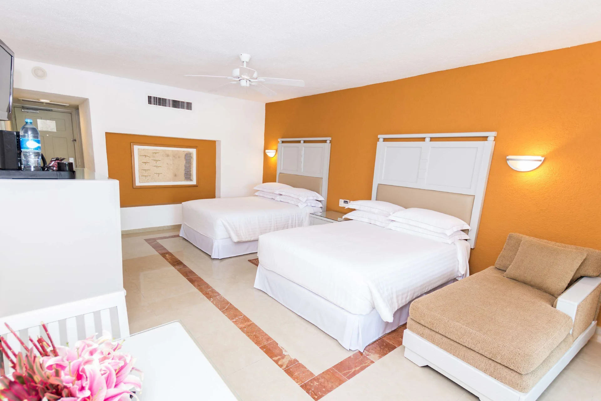 Two-bed guest room with chaise lounge and wall-mounted TV at Occidental Costa Cancún.