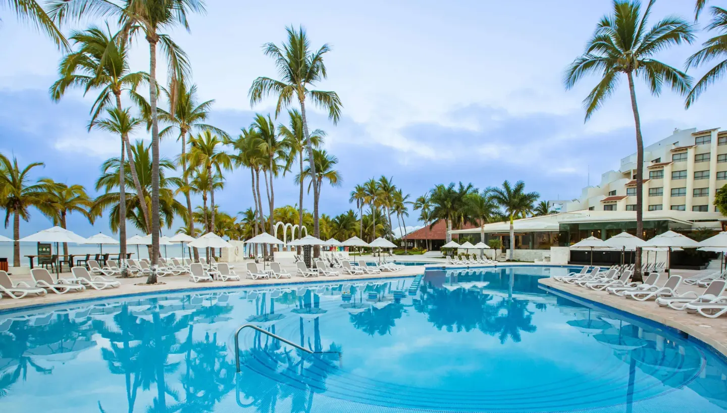 Resort pool with entry steps, palm trees, loungers, and ocean beyond at Occidental Nuevo Vallarta.