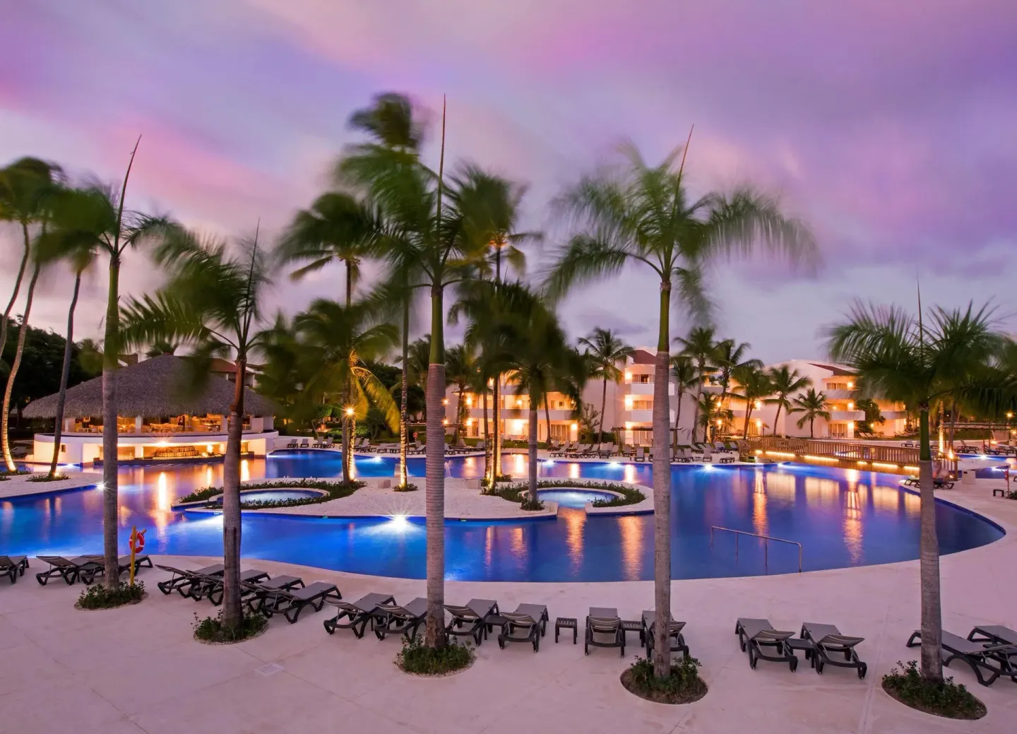 Large illuminated pool with palm islands at dusk at Occidental Punta Cana.