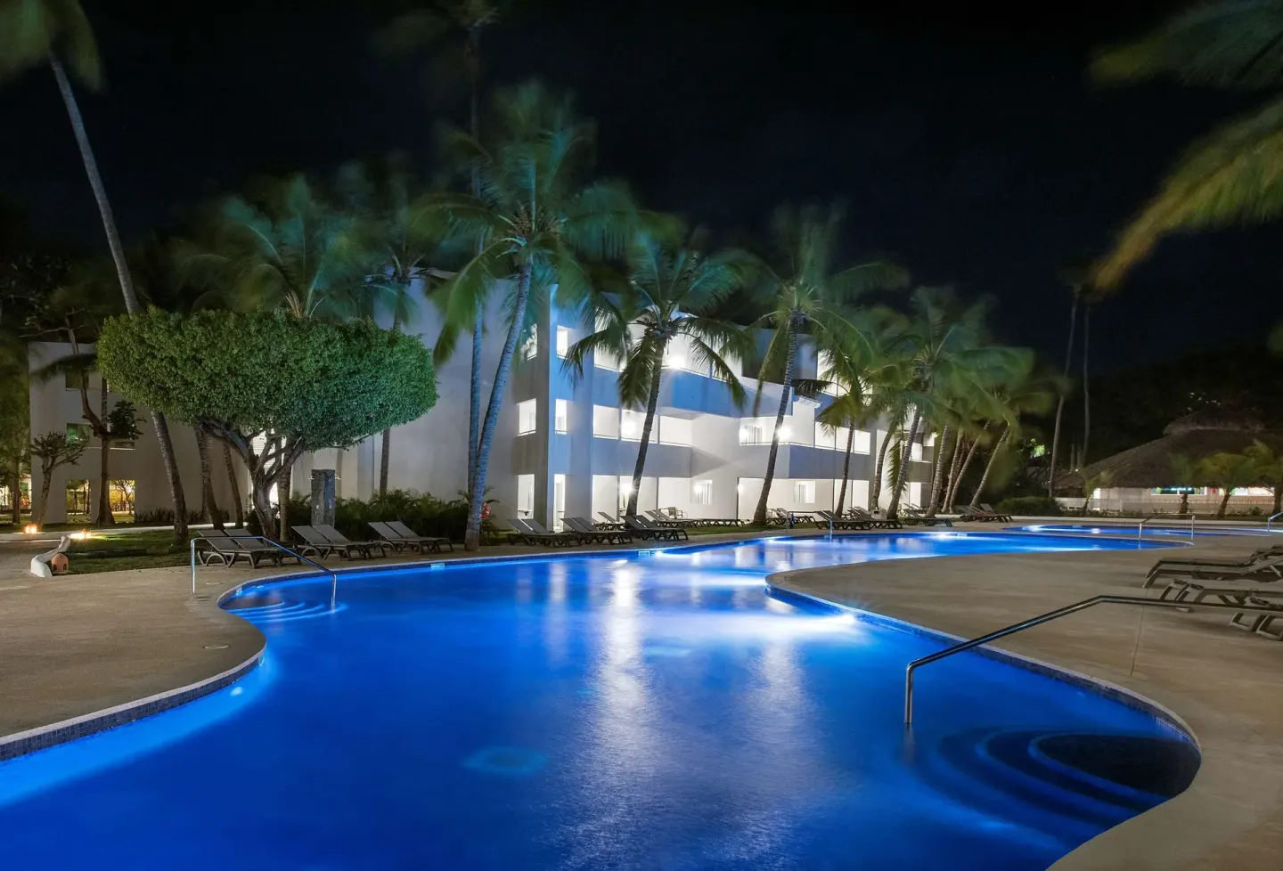 Night pool with blue lighting, palms, loungers, and guest building at Occidental Punta Cana.