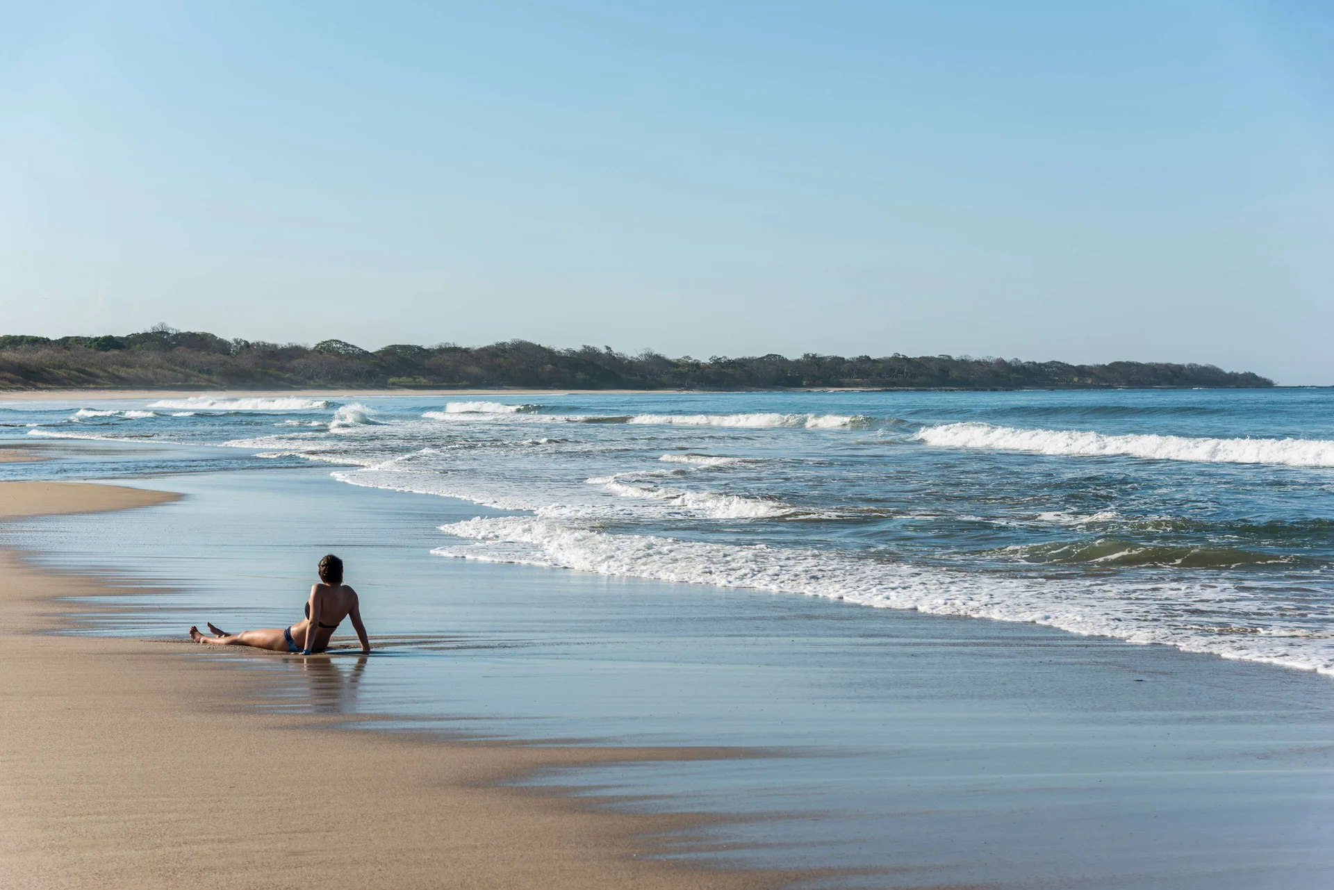 A beachgoer sits at the waterline on the sandy beach by Occidental Tamarindo.