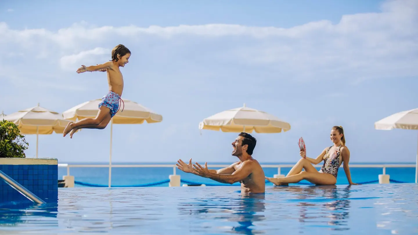 Family playing in the pool with ocean and umbrellas behind at Occidental Tucancún.