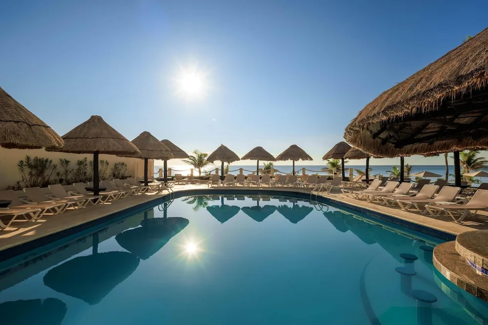 Pool with thatched shade structures and ocean beyond at Occidental Tucancún.
