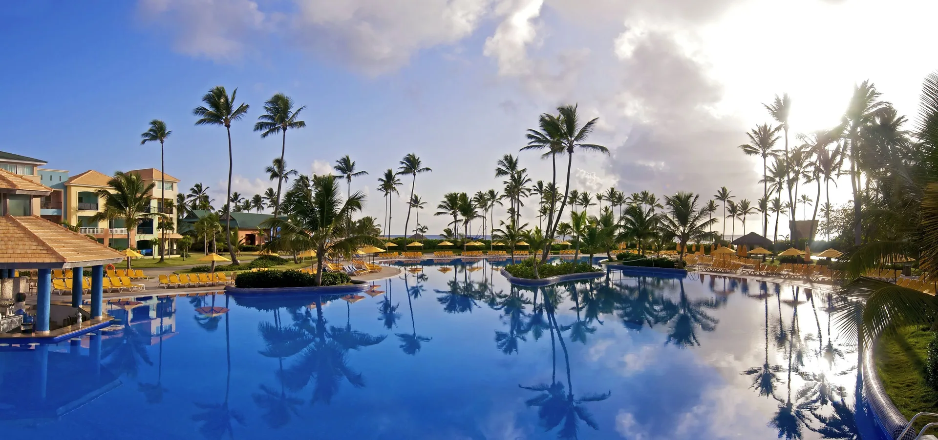 Sunlit pool with palm islands, yellow loungers and sea beyond at Ocean Blue & Sand Beach Resort.