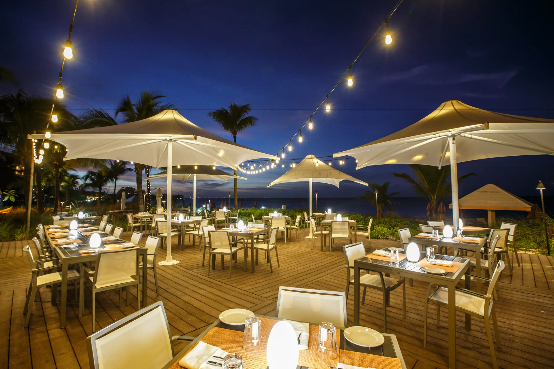 Beachfront dinner tables under umbrellas and string lights at Ocean Club West, Grace Bay Beach.