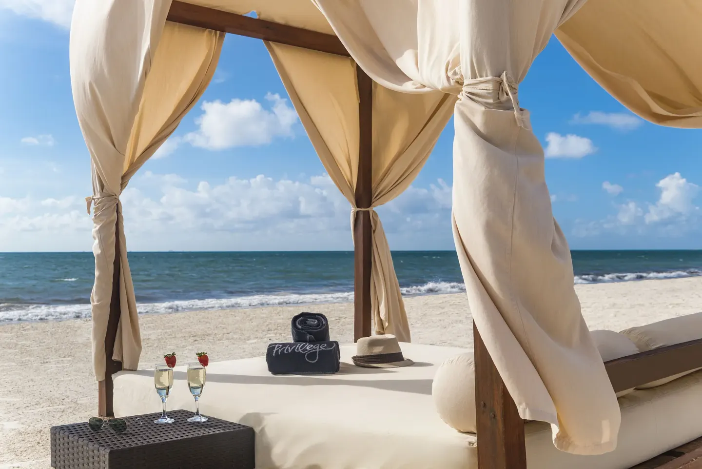 A curtained beach daybed with drinks and towels faces the ocean at Ocean Maya Royale.