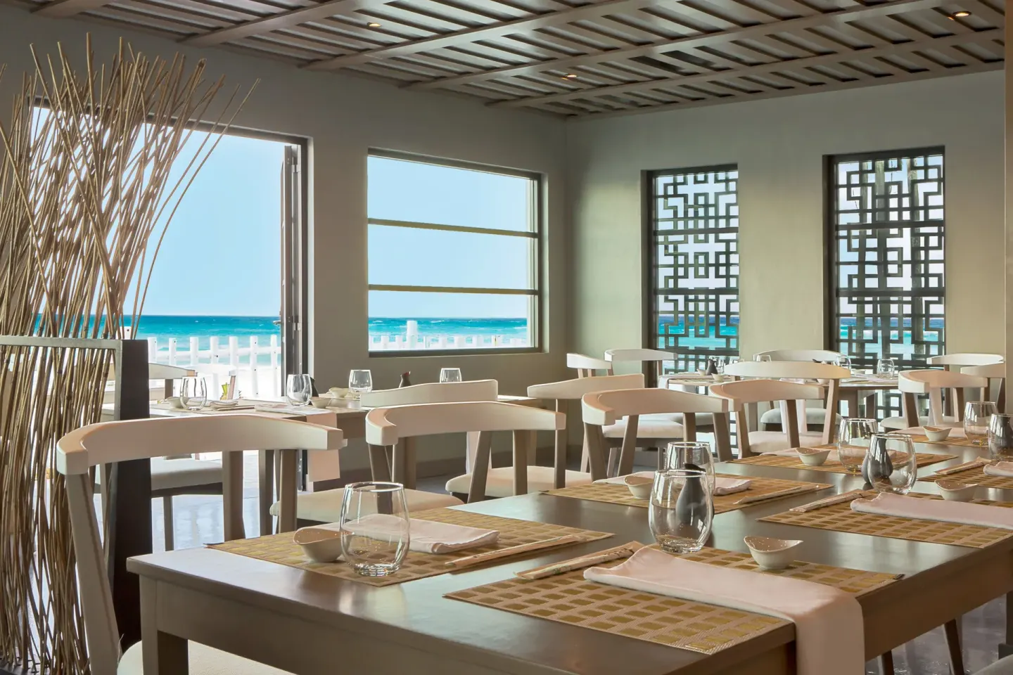 Ocean-view dining room with set tables and geometric screens at Oleo Cancun Playa Resort.