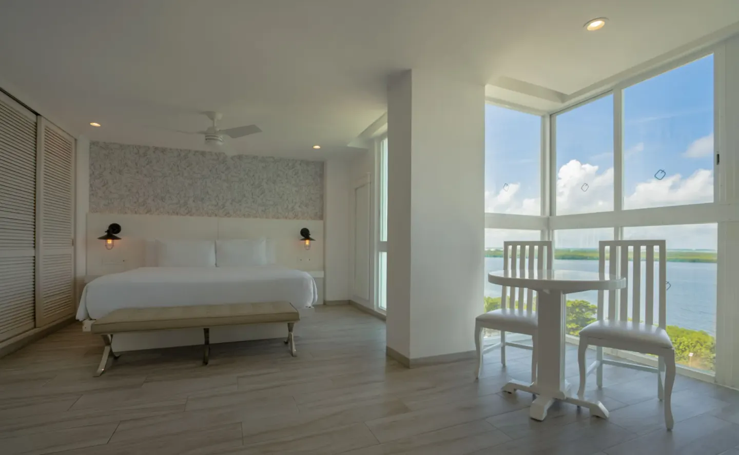 Junior Suite Sunset king bed and small table beside lagoon-view windows at Oleo Cancun Playa Resort.