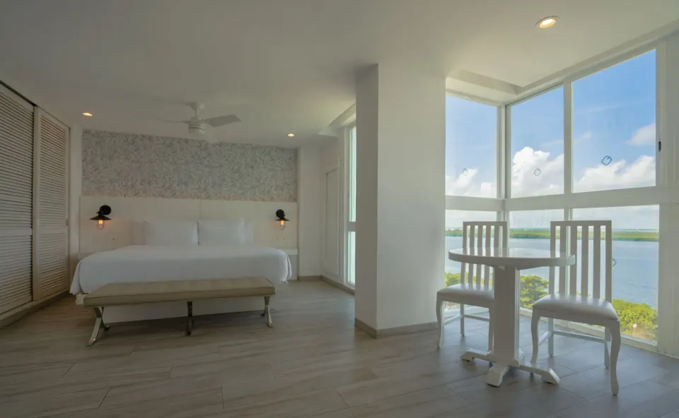 Junior Suite Sunset king bed and small table beside lagoon-view windows at Oleo Cancun Playa Resort.