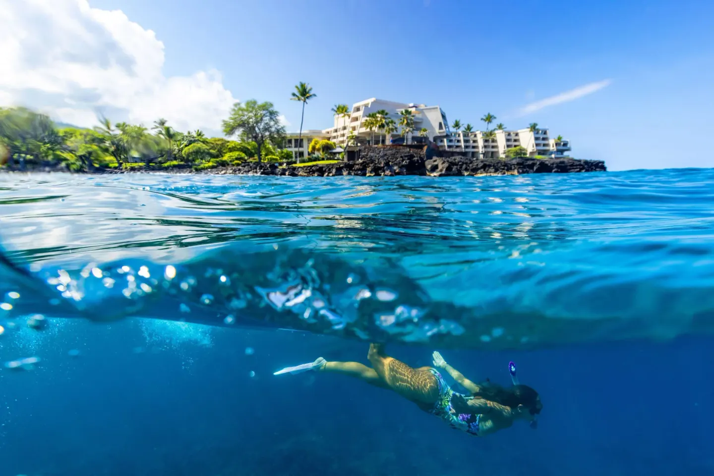 Snorkeler underwater with Outrigger Kona Resort and Spa visible on the shoreline.