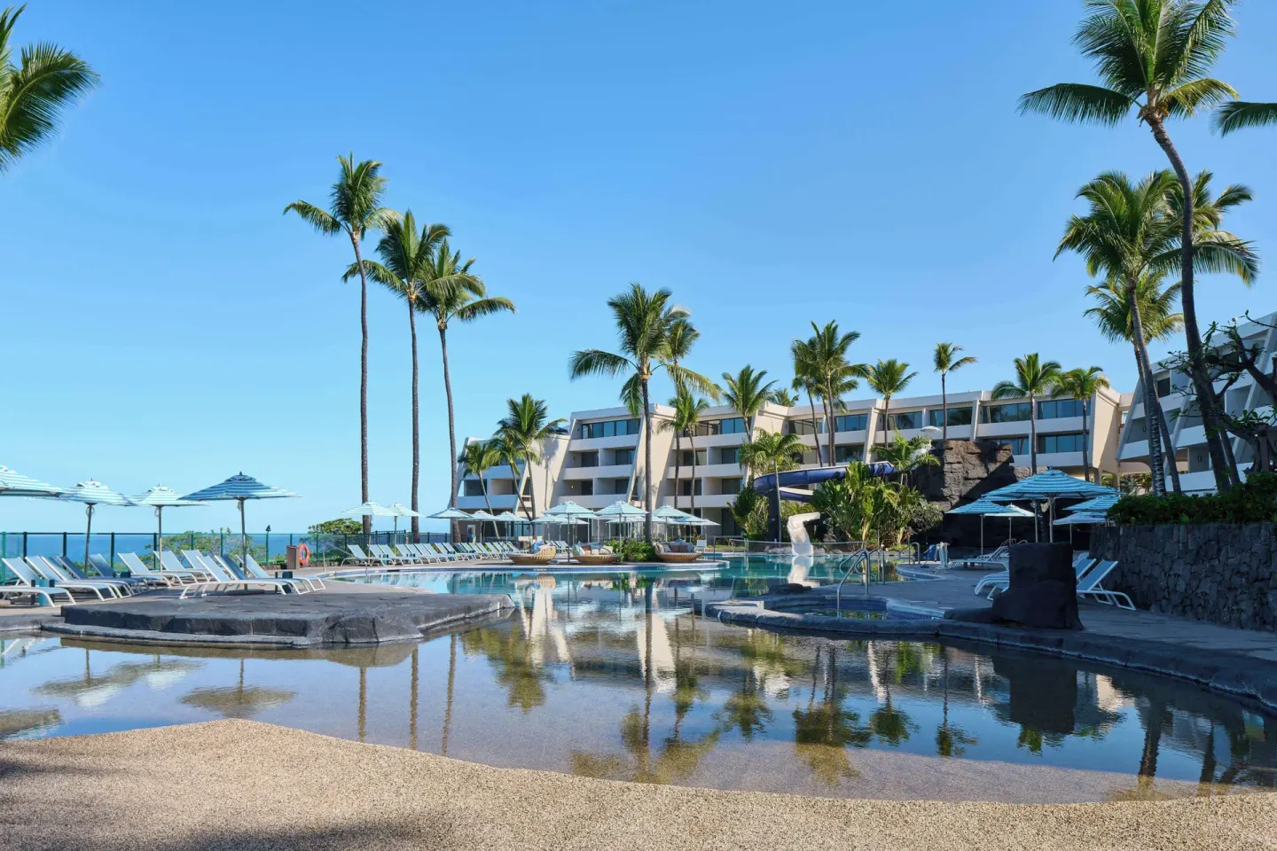 A wide resort pool scene with palms and guest buildings at Outrigger Kona Resort and Spa.
