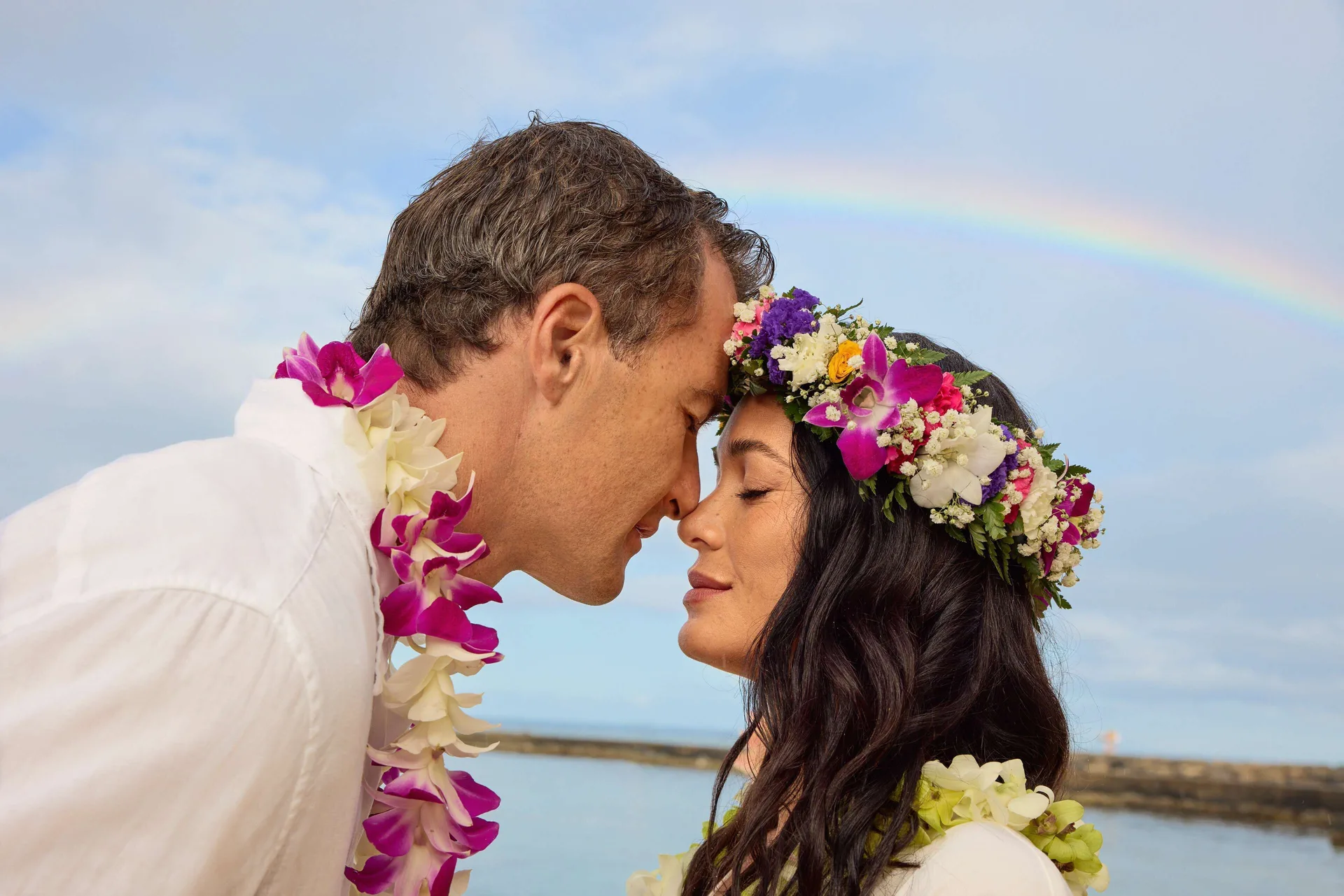 Couple wearing leis by the ocean and rainbow at OUTRIGGER Reef Waikiki Beach Resort.