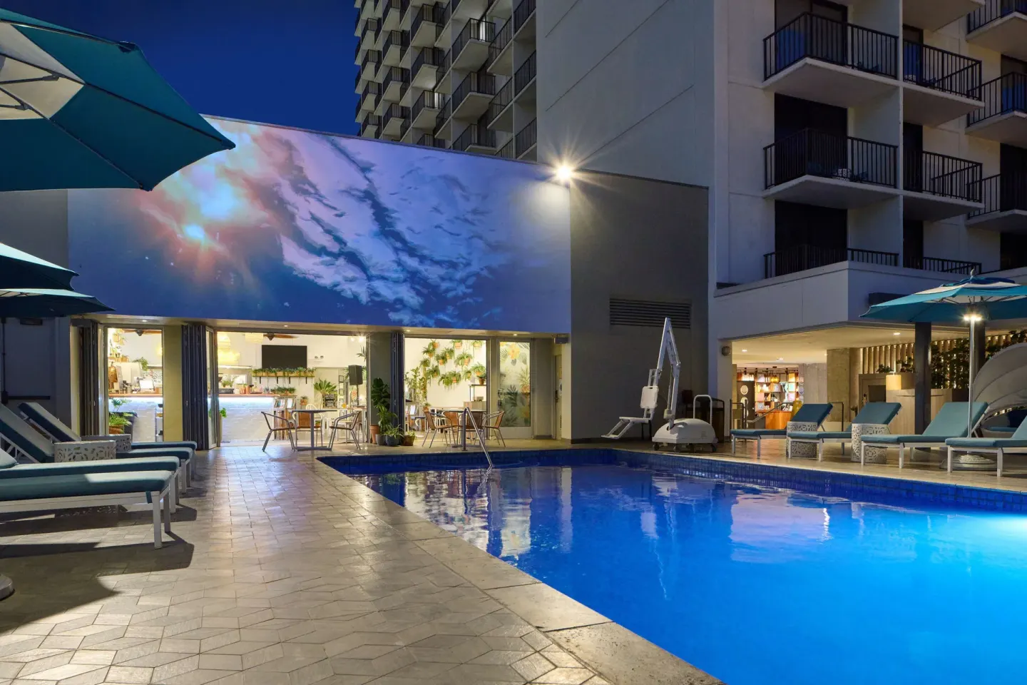 Evening pool scene with umbrellas and lit mural at OUTRIGGER Waikiki Beachcomber Hotel.