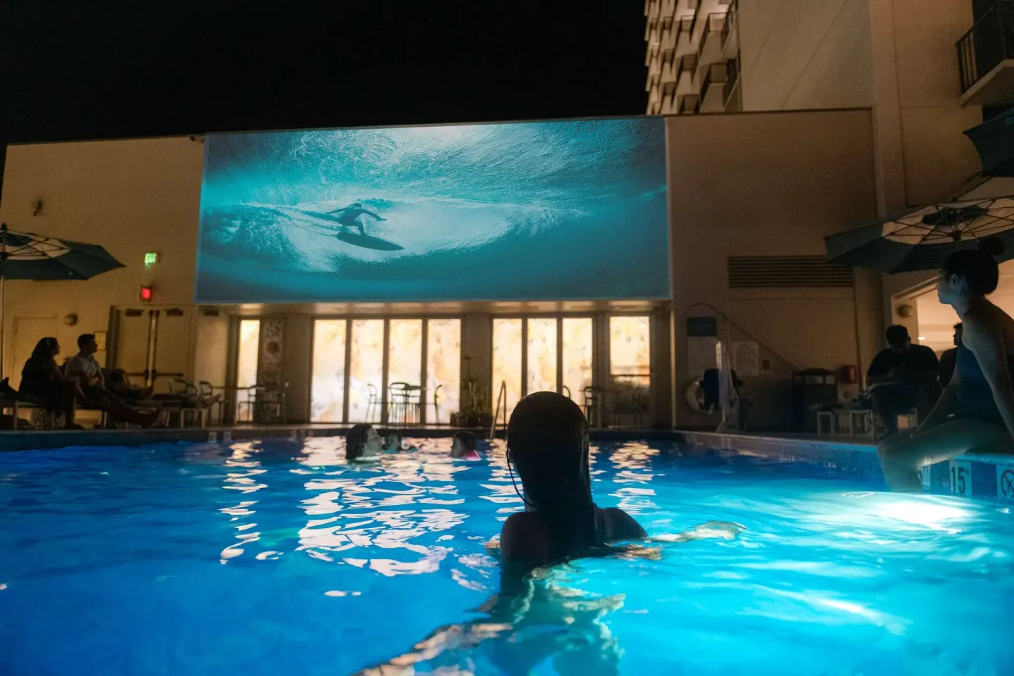 Nighttime pool with swimmers and a large surfing image at OUTRIGGER Waikiki Beachcomber Hotel.