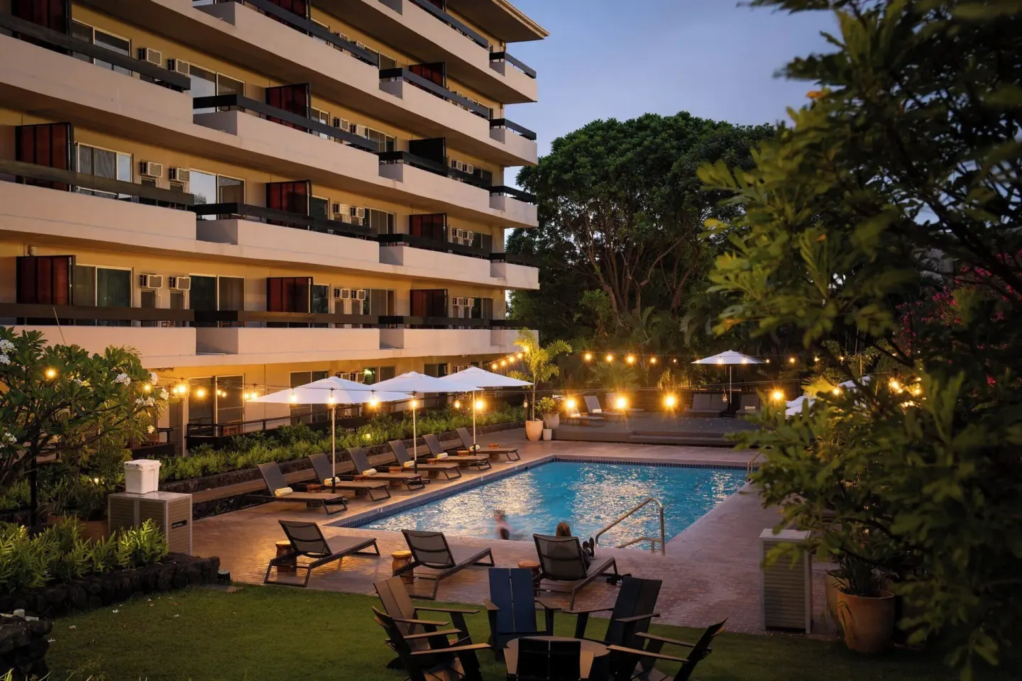 Evening pool courtyard with lit umbrellas, lounge chairs, and balconies at PACIFIC 19 Kona.