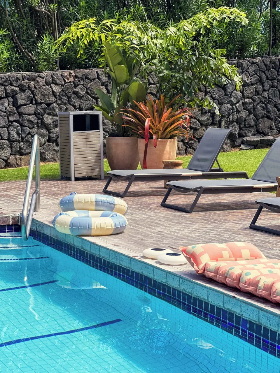 Pool edge with loungers, floats, potted plants, and a stone wall at PACIFIC 19 Kona.