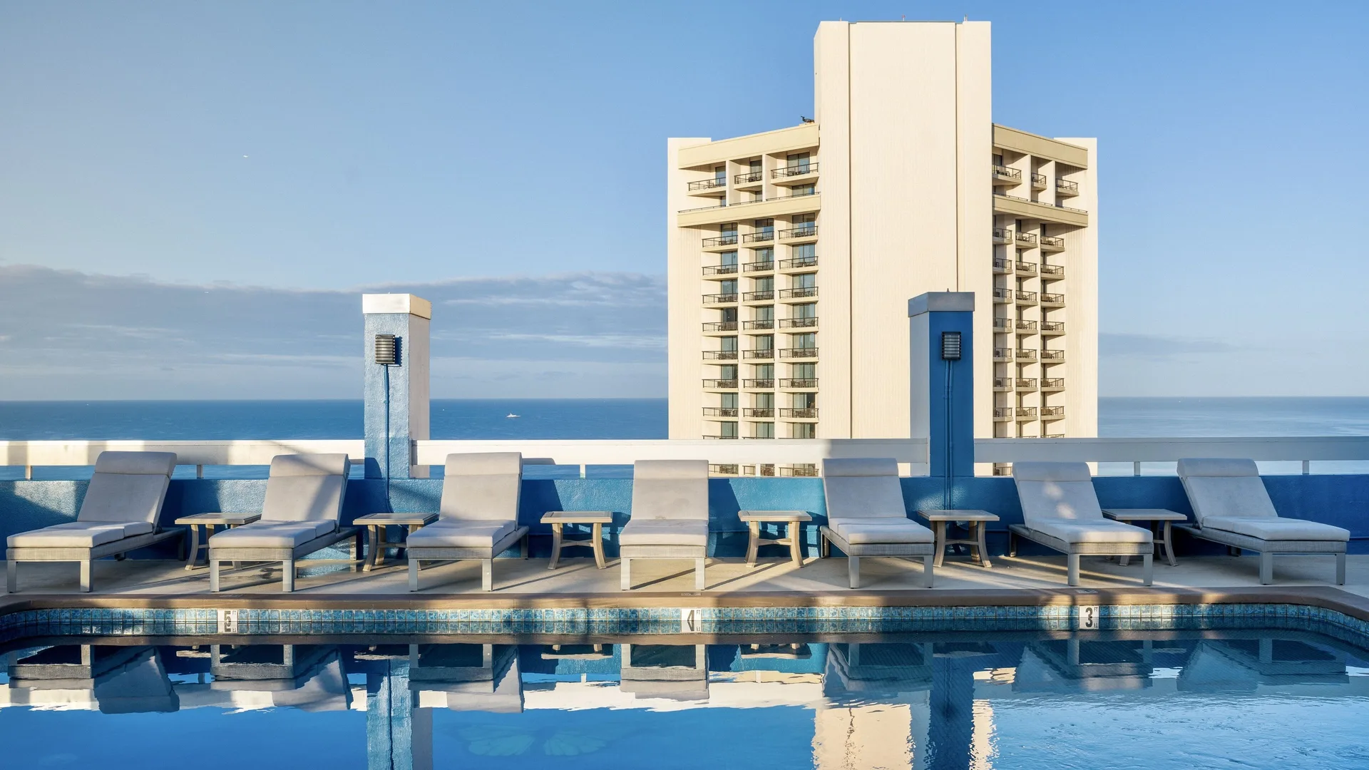 Pacific Monarch Hotel rooftop pool with loungers, blue railing, ocean, and tower view.