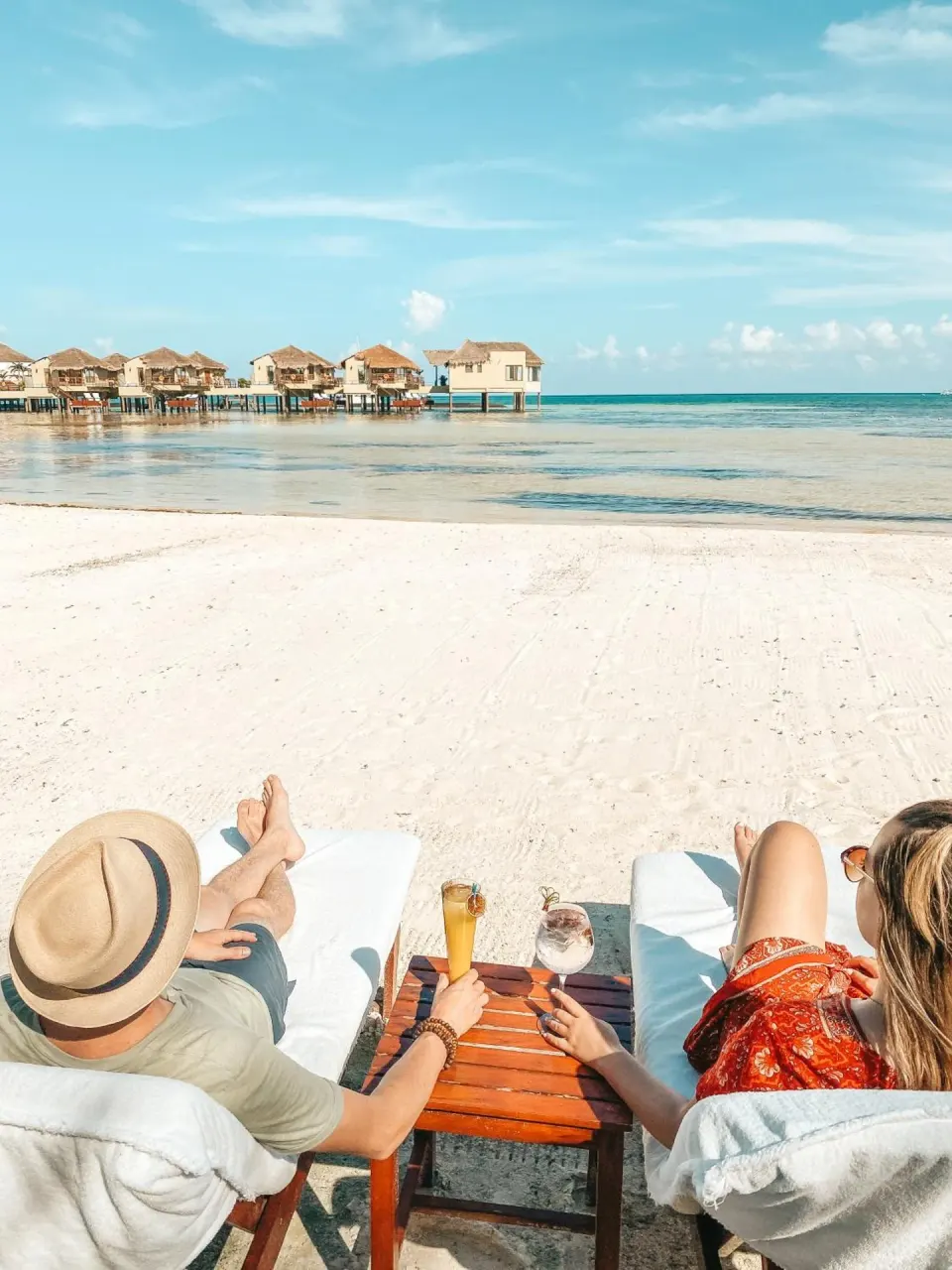 Couple relaxing on beach loungers facing overwater bungalows at Palafitos Overwater Bungalows.