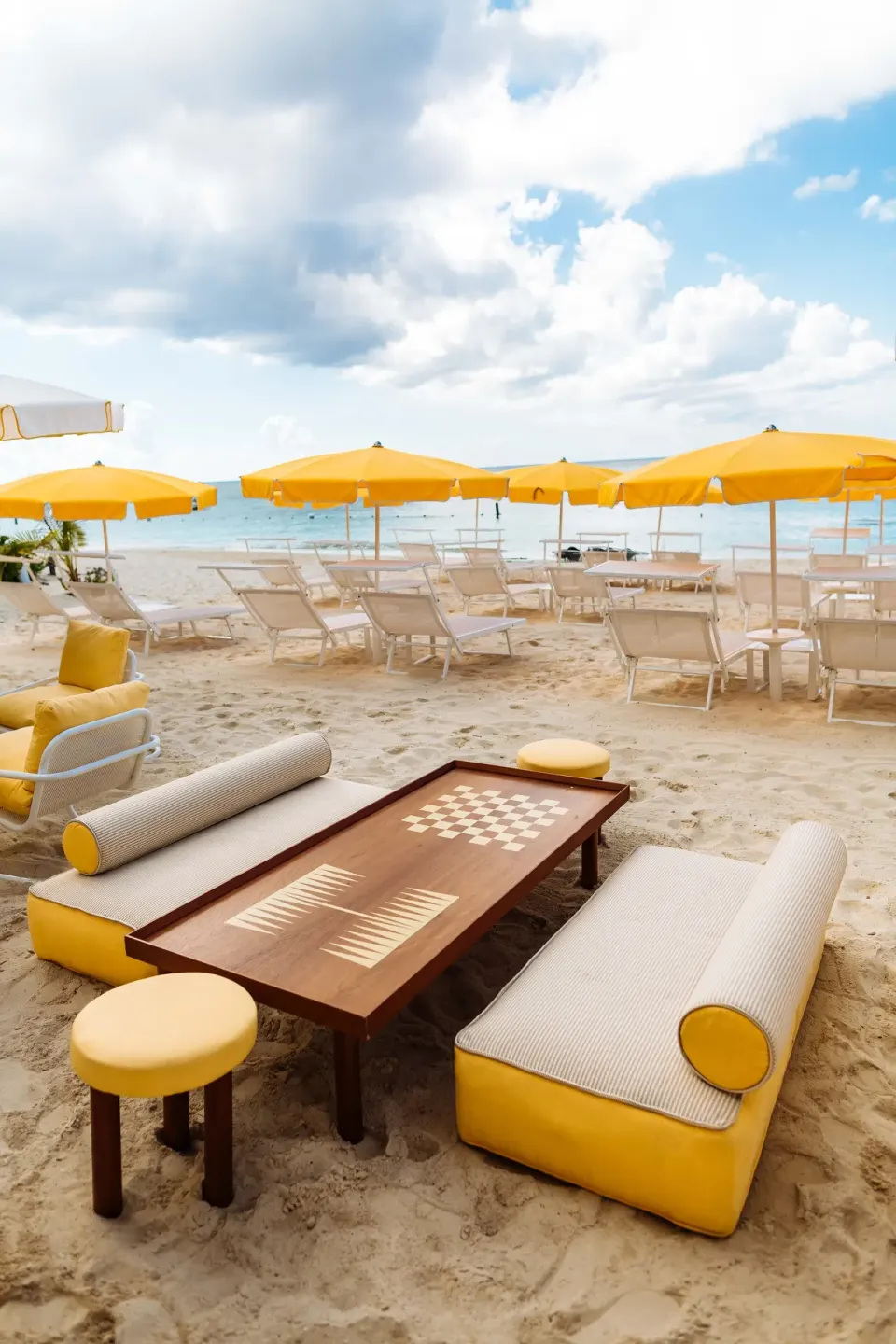 Low beach seating and a wooden game table with yellow cushions on the sand at Palm Heights.