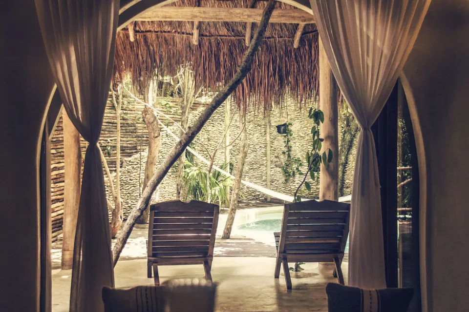 Shaded room opening to a thatched patio with two lounge chairs and a pool at Papaya Playa Project.