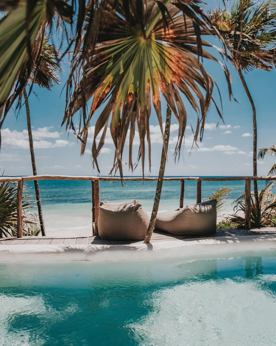 Pool edge and two beanbag loungers beneath palms with beach beyond at Papaya Playa Project