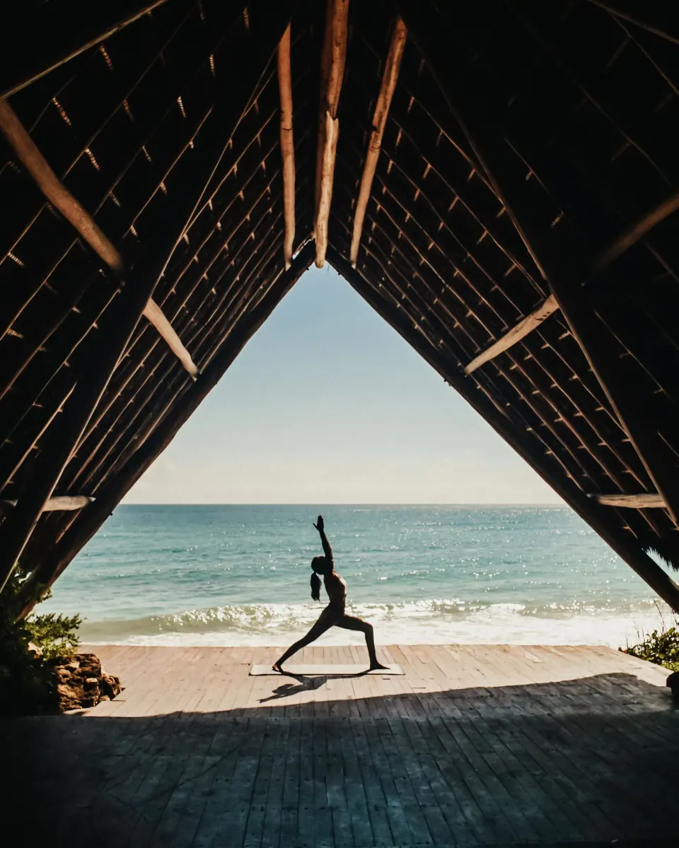 Silhouetted person practicing yoga under a thatched palapa facing the ocean at Papaya Playa Project