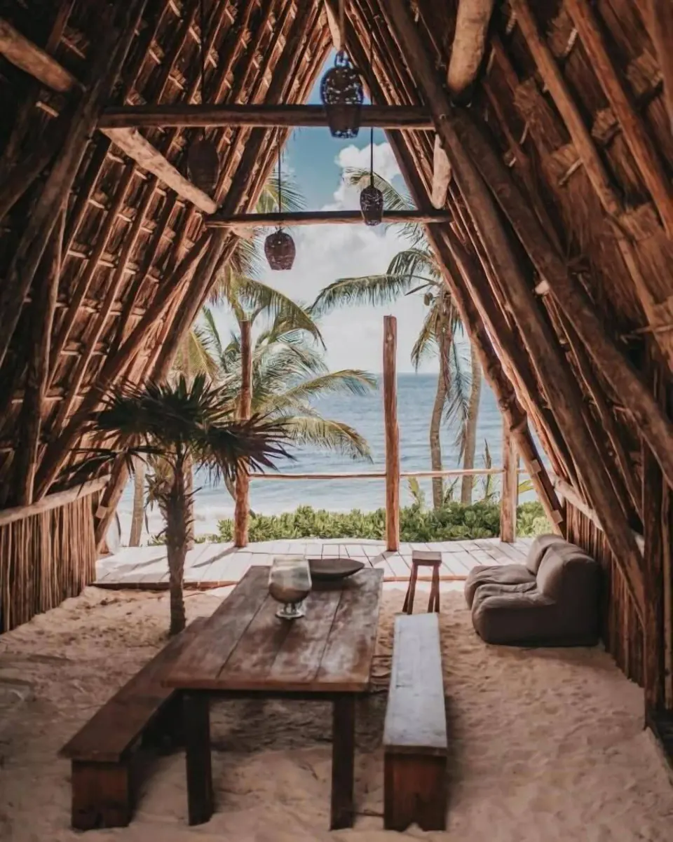 View from inside Casa Arena palapa toward the beach with wooden table and sand floor at Papaya Playa Project