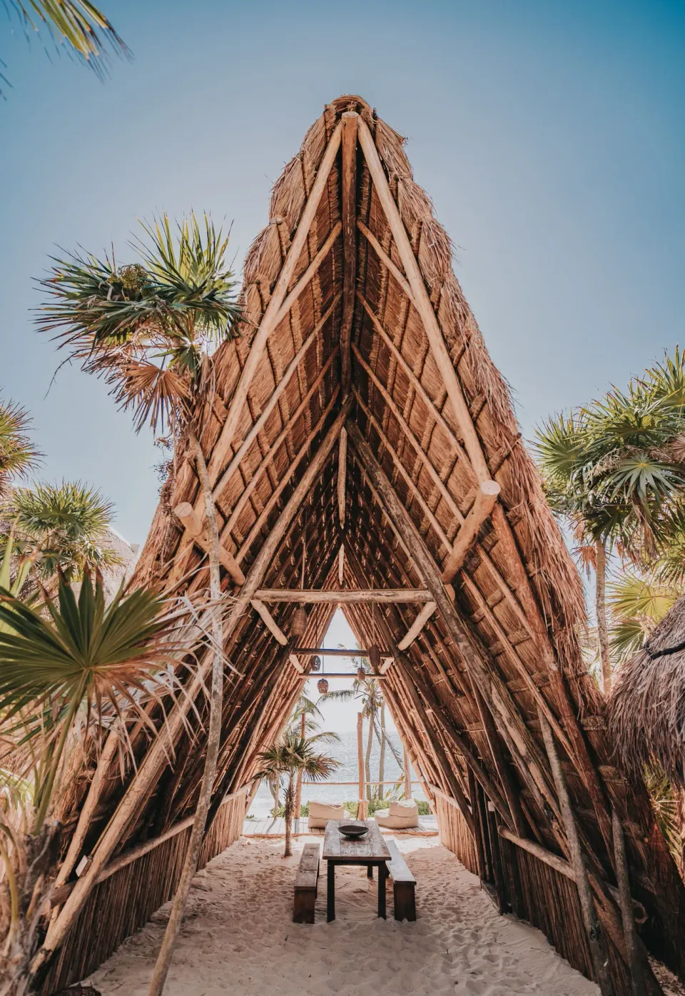 Tall A-frame palapa structure with a communal table leading toward the beach at Papaya Playa Project