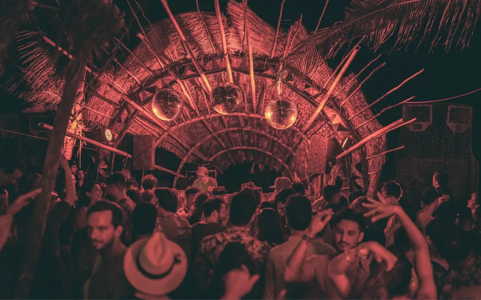 Nighttime crowd dancing under a thatched arch DJ booth at Papaya Playa Project